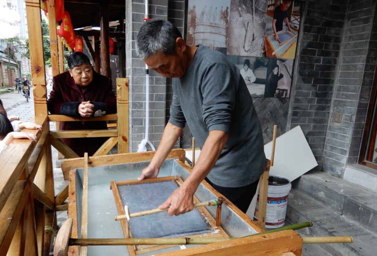 The papermaking craftsman in the image skillfully holds a bamboo screen, expertly scooping up wet paper from the pulp pool.