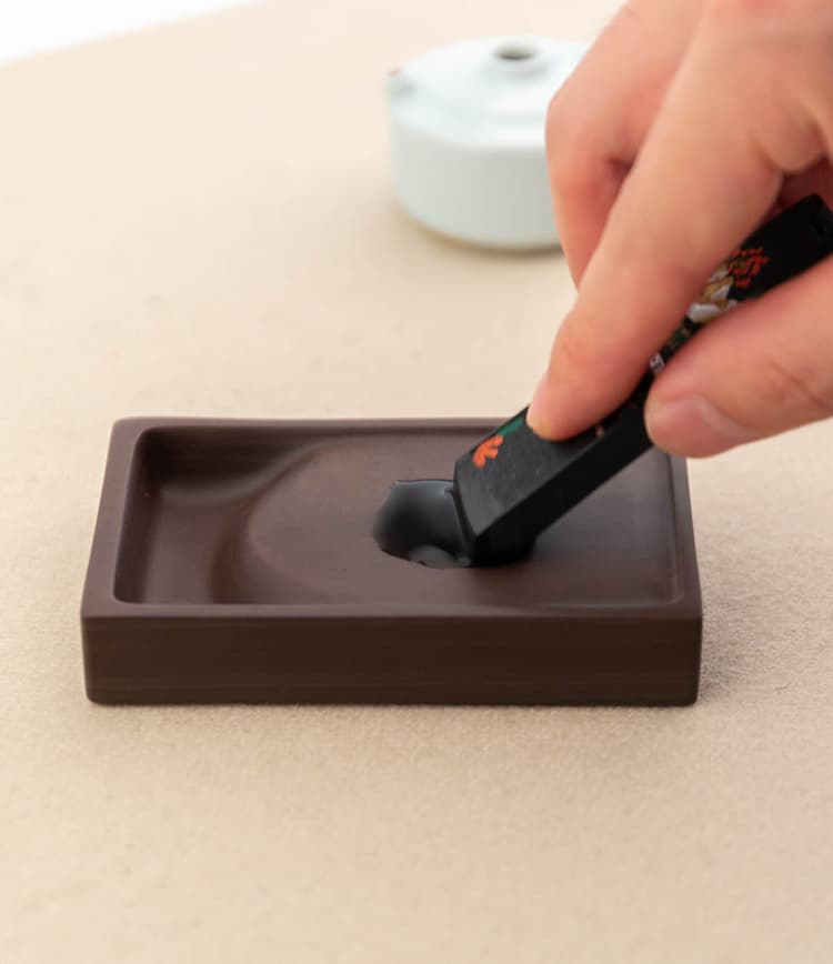 You add an appropriate amount of water to an inkstone, take an ink stick, and grind it back and forth on the inkstone. 